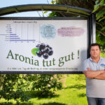 Mann steht vor Infotafel „Aronia tut gut“ mit Diagramm zu Vitaminen und Anthocyanen – Symbol für Erfahrung und Beratung im Aronia-Anbau